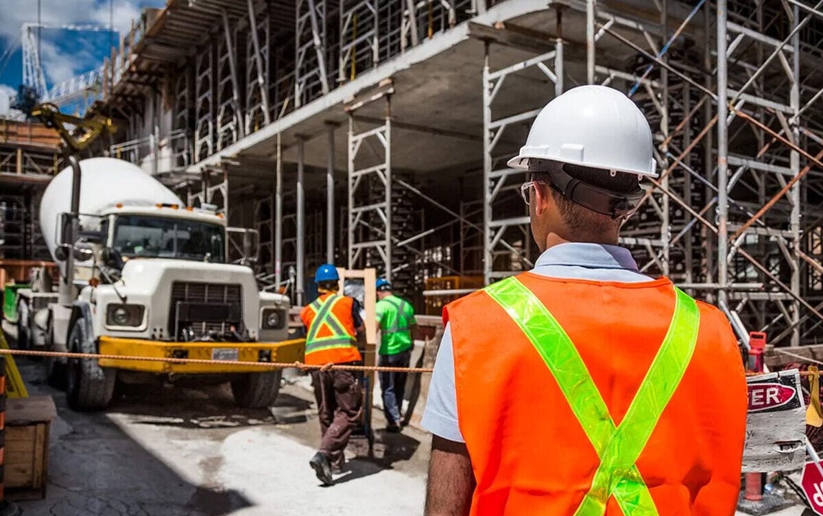 Man on construction site in white construction hat with truck and two men in the background